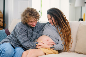 Obraz premium Husband touching pregnant tummy of his charming joyful wife which lying on the couch in cozy living room