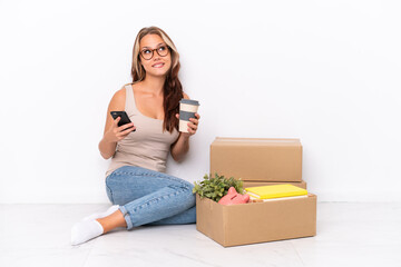 Young Russian girl sitting on the floor isolated on white background holding coffee to take away and a mobile while thinking something