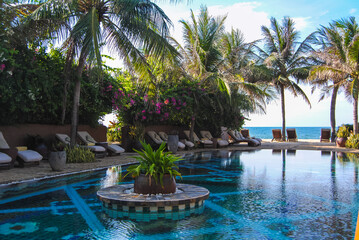 A view of pool, palm trees and comfortable lounge chairs in the shade of plants and flowers in a luxurious hotel