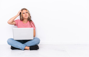 Young Russian girl with a laptop sitting on the floor isolated on white background having doubts...