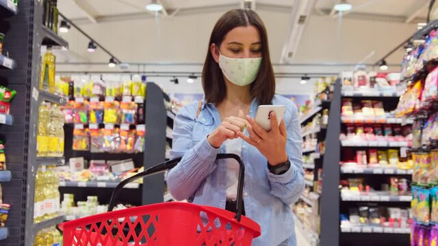 Close up video of a young woman in a protective mask holding the grocery basket and using her cellphone in modern market to follow the list of products