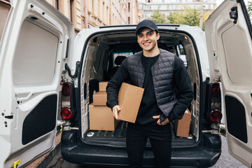 Portrait of a smiling courier holding a cardboard box while standing at car trunk looking at camera