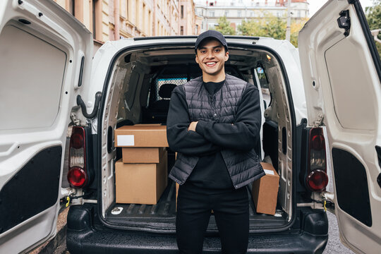Portrait of a smiling delivery guy wearing a cap and uniform standing with crossed arms at van trunk looking at camera