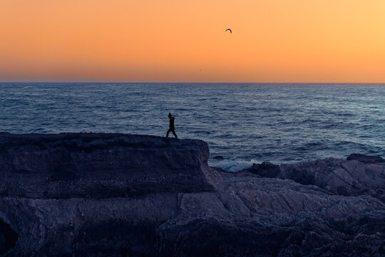 Sunset Seascape. Rocky Cliffs And Silhouette Of Walking Woman, Blue Ocean And Bright Yellow Sky On Background