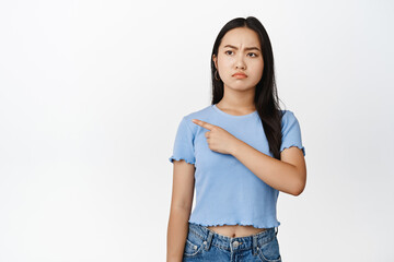 Angry asian girl pointing, looking left with grumpy frowning face, stare with dislike, standing in tshirt over white background