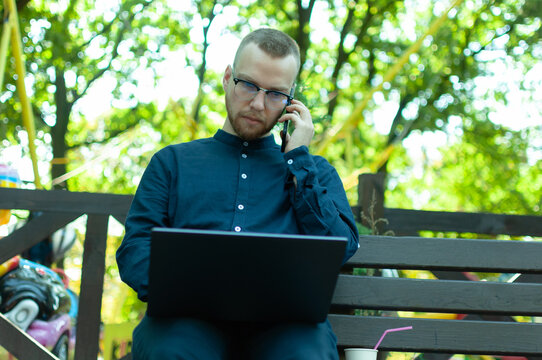 Young Caucasian male working on a laptop and talking on the phone in a park