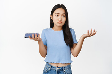 Confused asian girl holding smartphone and shrugging shoulders indecisive, dont know, cant understand, standing over white background