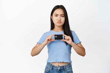 Angry asian girl showing credit discount card, frowning and looking disappointed at camera, standing over white background