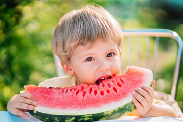 Lovely toddler boy eating fresh red watermelon. Yummy portrait of cheerful child sits by the table...