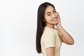 Beauty and skincare. Young asian woman touches her facial skin, smiling and gazing at camera, standing in yellow t-shirt over white background