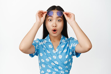 Portrait of asian girl takes off sunglasses and looking amazed at smth awesome, checking out promo deal, standing over white background