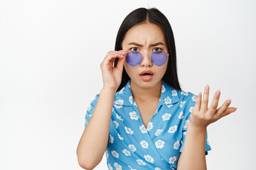 Close up portrait of asian girl in sunglasses looking confused and disappointed, cant understand smth, standing over white background