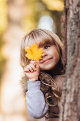 Beautiful blonde girl covers one eye with yellow maple leaf outdoors in autumn park.