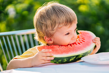 Handsome toddler boy eating fresh red watermelon. Yummy portrait of smiling child sits by the table in garden. Healthy lifestyle, melon vitamins, summer harvest.