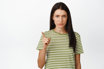 Fototapeta premium Angry brunette woman shaking finger in disapproval, scolding bad behaviour, standing over white background in t-shirt