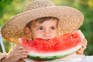 Cute little boy eating slice of juicy watermelon sitting on natural green garden background. Adorable kid in straw hat enjoying summer fresh melon fruit with smile.