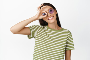 Happy smiling brunette girl in sunglasses, showing okay, zero gesture, standing in summer clothes...