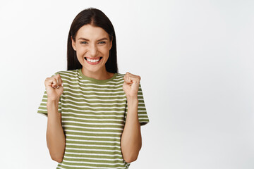 Excited brunette woman achieve goal, cheering and celebrating, winning prize, standing in t-shirt over white background