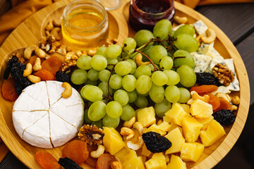 cheeses with grapes, dried fruits, nuts, honey and jam on a round wooden plate.