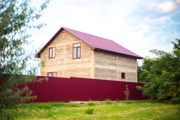 Fototapeta na wymiar A house with a crimson roof behind a fence in a village in summer. Construction of suburban housing