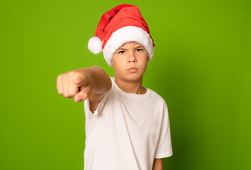 Portrait of a serious boy in a red hat and white t-shirt pointing finger to camera on a green background. A child in a Christmas hat of Santa Claus.