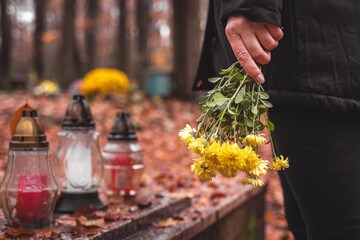 Paying respect for dead person. Mourning woman holding flowers in hands and standing at grave in cemetery. 