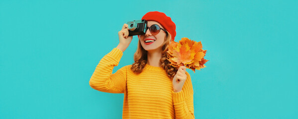 Autumn portrait of happy smiling young woman with film camera and yellow maple leaves wearing a sweater, french beret on blue background