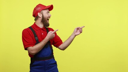 Excited positive worker man points fingers aside, showing copy space for advertisement or promotion, wearing blue uniform, red T-shirt and visor cap. Indoor studio shot isolated over yellow background