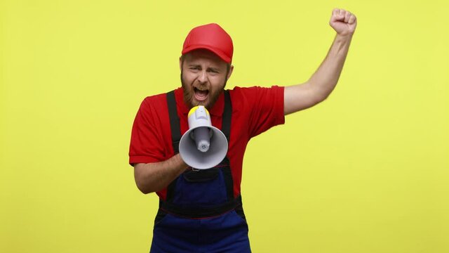 Angry bossy workman shouting in megaphone with aggressive facial expression, raised arm, protesting, wearing blue uniform, red T-shirt and visor cap. Indoor studio shot isolated over yellow background