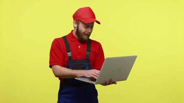 Confused workman holding notebook in hands and expressing negative emotions, screaming why, wearing blue uniform, red T-shirt and visor cap. Indoor studio shot isolated over yellow background.