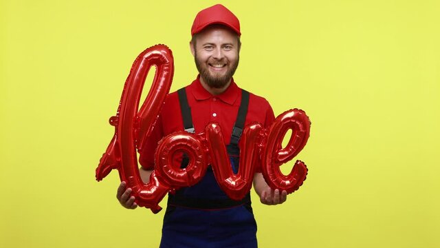 Extremely happy courier man delivering air balloons to clint, holding foil love word in hands, wearing blue uniform, red T-shirt and visor cap. Indoor studio shot isolated over yellow background.