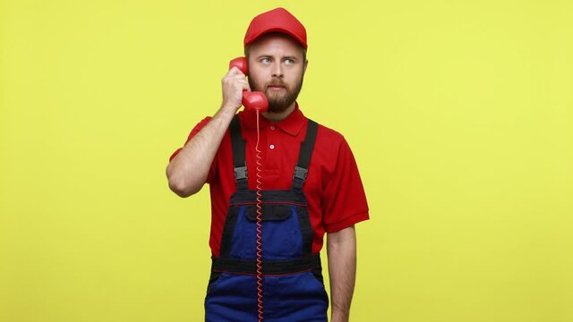 Portrait of angry call center worker screaming on his client, expressive aggressive emotions, wearing blue uniform, red T-shirt and visor cap. Indoor studio shot isolated over yellow background.