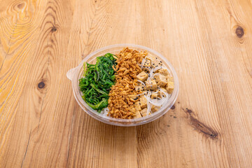 Vegan poke bowl with tofu, chopped white onion, wakame seaweed salad, crispy fried onion, sesame and poppy seeds with white rice base in take away container
