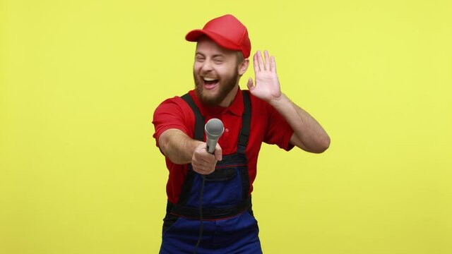 Positive handy man having fun on corporate party, offering microfone to colleague to sing with him, wearing blue uniform, T-shirt and visor cap. Indoor studio shot isolated over yellow background.