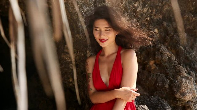 Alex Portrait of pretty young woman in red dress standing in front of rock, posing. Girl looking at camera and smiling shyly. Woman with bright makeup matching dress colour making picture during