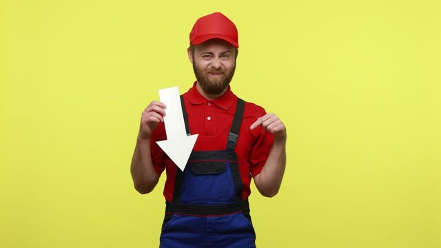 Upset disappointed master holding white paper arrow pointing down, being sad of decrease, wearing blue uniform, red T-shirt and visor cap. Indoor studio shot isolated over yellow background.