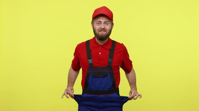 Handsome workman showing his empty porckets, expressing sadness, has no orders and salary, wearing blue uniform, red T-shirt and visor cap. Indoor studio shot isolated over yellow background.