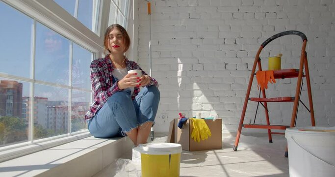 Young caucasian woman decorating wall doing house renovation. A female painter taking a break on the floor in front of a painted wall in an apartment. Home improvement concept. New home concept. 4k sl