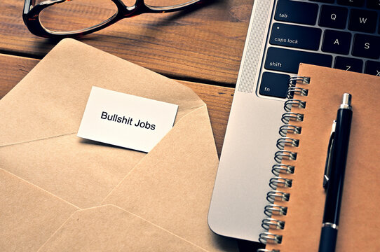 On the table, there is a card with the word of Bullshit Jobs along with a laptop computer and glasses.