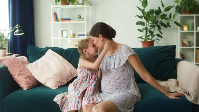 Happy pregnant mother and her daughter sitting on sofa in living room, hugging, touching tummy. Portrait of woman expecting child, smiling and stroking belly. Pregnancy and motherhood concept.