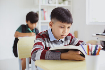 Serious Asian boy reading a book during a lesson in a classroom
