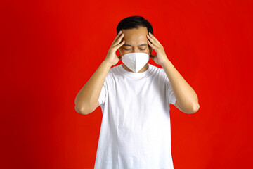 Asian man wearing a medical mask, having a headache while holding his head with both hands on a red background