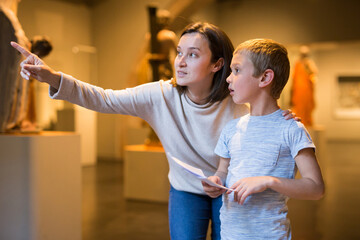 Young female tutor with boy looking at exposition in museum of ancient sculpture, pointing to...