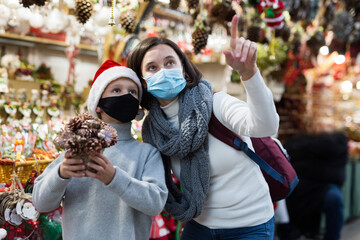 Happy tween boy with his mother in protective masks to prevent spread of viral infections choosing Xmas decorations and gifts on street fair ..