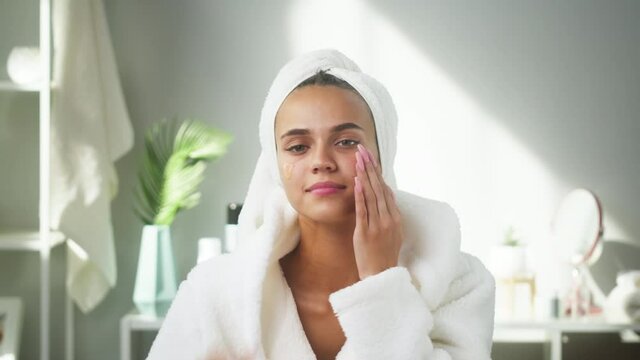 Woman applying face serum using pipette for moisturizing, morning skincare routine. Young female student wearing bathrobe looking in mirror in spa salon. Natural day make up concept.