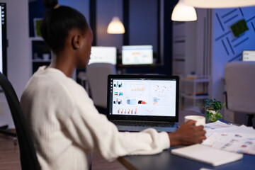 African american business entrepreneur analyzing financial company graphs on laptop computer overworking at annual reports late at night in startup office. Manager typing marketing statistics