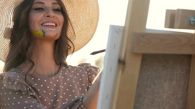 Close-up Artist in summer dress and big hat stands on mown field in rays of setting sun and draws with brushes on canvas standing on an easel holding brushes in her teeth. Painter draws in nature.