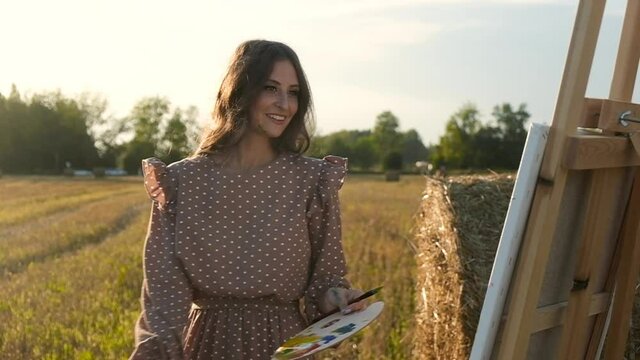 Happy young artist, stained with paints, stands on a field with haystacks in the rays of the setting sun and cheerfully draws on canvas on an easel with brushes. Painter draws in nature.