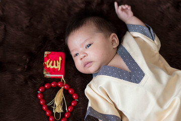 A cute Asian infant Muslim in thobe and rosary beads on red, muslim bead on bed in the home, translation is the name of muslim God "Allah" in arabic text