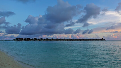 A row of water villas stretches over the aquamarine ocean. Small waves roll on the sandy beach....